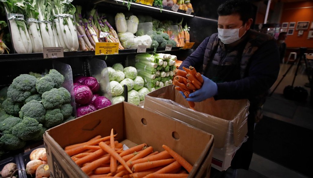 In this March 27, 2020, file photo, a worker, wearing a protective mask against the coronavirus, stocks produce before the opening of Gus's Community Market in San Francisco. (AP Photo/Ben Margot, File)